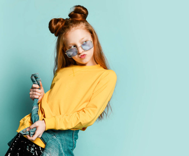 Cool red-haired teen girl poses in blue smoked glasses, yellow sweatshirt, glossy black skirt is making kissy lips - Foto, Imagem