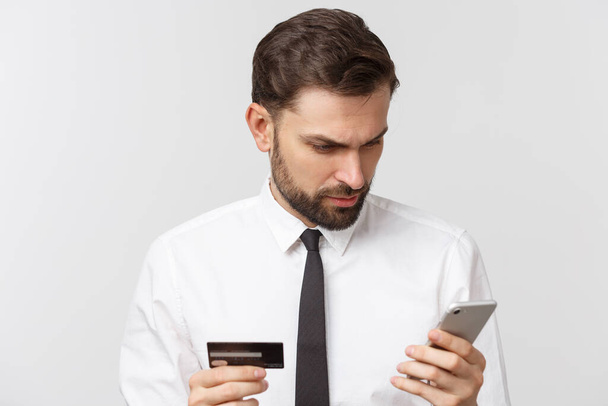 Zdjęcie pewnego przedsiębiorcy człowieka w garniturze i krawat trzymając smartfon i kartę kredytową do płacenia online izolowane na szarym tle. - Zdjęcie, obraz