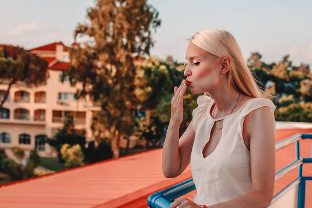 Beautiful stylish blonde woman in fashionable clothes posing on the balcony and blows a kiss. - Photo, Image