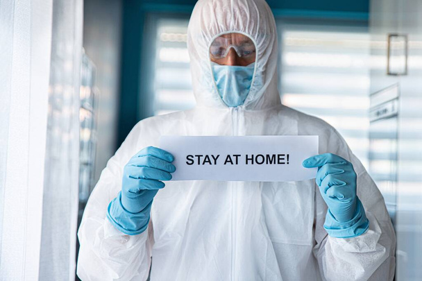Mann in Anti-Virus-Schutzanzug und Maske hält Karte mit Botschaft zu Hause - Foto, Bild