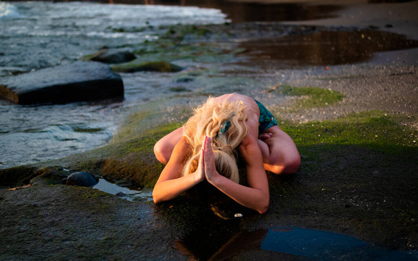 Joogaharjoitukset. Blondi joogi harjoitellaan Lotus poseeraa takana mutka. Kädet namumaisessa mudassa. Itsehoidon käsite. Joogaretriitti. Tanah Lotin ranta, Bali, Indonesia
 - Valokuva, kuva