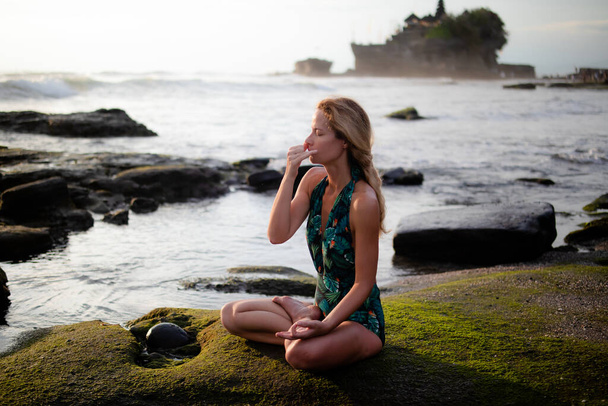 Młoda kobieta siedząca w pozycji lotosu, praktykująca Anuloma Viloma Pranayama, Alternate Nostril Breathing. Kontroluj ranę poprzez kontrolę oddechu. Ćwiczenia oddechowe. Odwrót jogi. Tanah Lot plaża, Bali - Zdjęcie, obraz