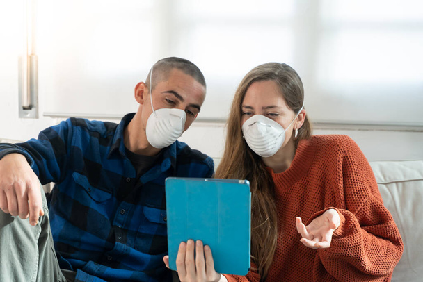 Coronavirus. A couple sitting on a sofa on a quarantine at home watching tv. Couple at home on the couch, both wear masks because of avoiding contagion. Stay at home.  - Fotografie, Obrázek