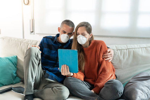 Coronavirus. A couple sitting on a sofa on a quarantine at home watching tv. Couple at home on the couch, both wear masks because of avoiding contagion. Stay at home.  - Foto, immagini