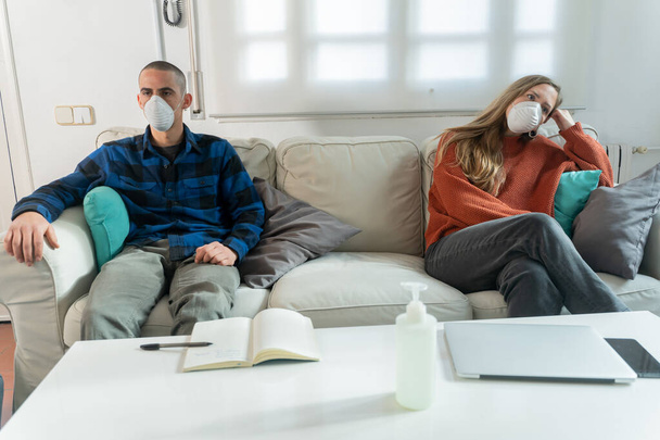 Coronavirus. Un couple assis sur un canapé en quarantaine à la maison regardant la télévision. Couple à la maison sur le canapé, les deux portent des masques en raison d'éviter la contagion. Reste à la maison.
.  - Photo, image
