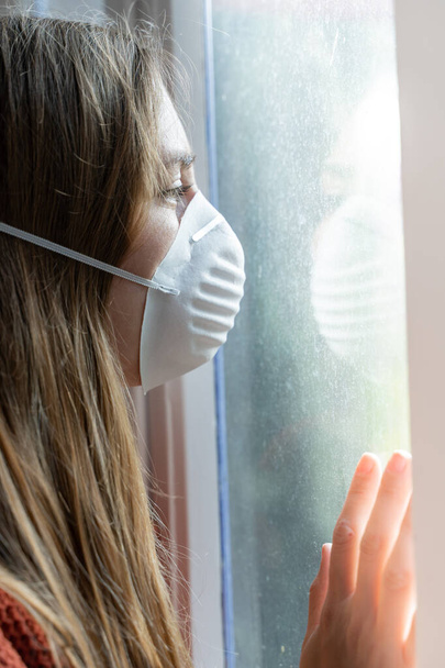 Ιός του κερατοειδούς. Άρρωστη γυναίκα από τον ιό Κορόνα που κοιτάζει από το παράθυρο και φοράει μάσκα προστασίας και ανάρρωσης από την ασθένεια στο σπίτι. Καραντίνα. Ασθενής απομονωμένος για πρόληψη μόλυνσης. Σπίτι. - Φωτογραφία, εικόνα