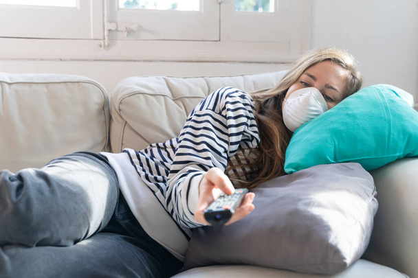 Coronavirus. Woman at home wearing protective mask. Woman in quarantine for coronavirus on the couch cleaning her hands with sanitizer gel. Working from home. Clean your hands with sanitizer gel.  - 写真・画像