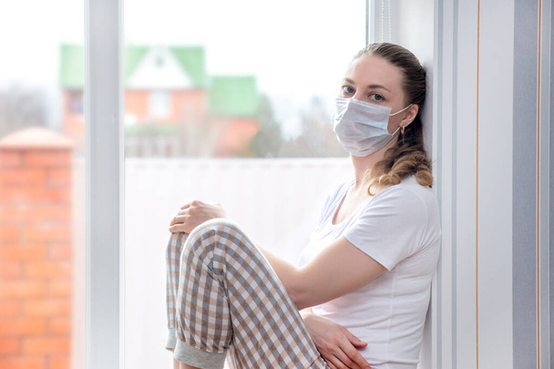 Home quarantine. Caucasian woman on sick leave is sitting at window in a medical protective mask, looking sad. Poor health, a seasonal epidemic of the flu virus and colds. Home treatment - Фото, изображение
