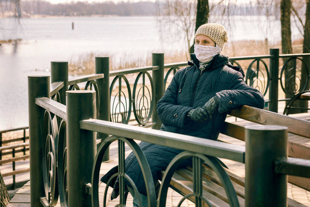 Chudá starší žena nosí domácí masku, aby se chránila před viry, jako je koronavirus, také známý jako covid-19, nebo SARS a MERS. Sedí na lavičce u řeky. - Fotografie, Obrázek