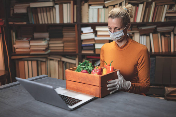 Woman with protective mask from viruses and pathogens using laptop at home, oreding food, groceries, supplies over the internet. - Photo, Image