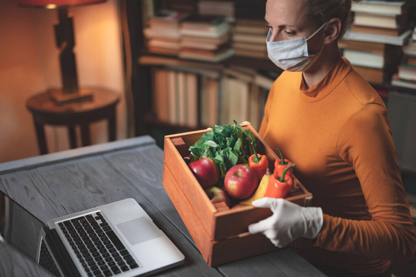 Kobieta z maską ochronną przed wirusami i patogenami korzystająca z laptopa w domu, z jedzeniem, artykułami spożywczymi, zapasami przez internet. - Zdjęcie, obraz