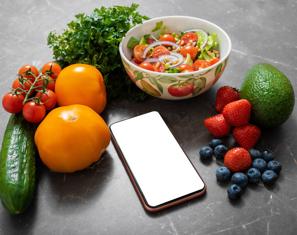 野菜や果物に囲まれたキッチンテーブルの上に白い空の画面を持つスマートフォン - 写真・画像