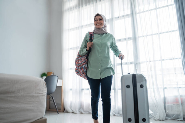 smiling young hijab traveler carrying her bag and holding suitcase - 写真・画像