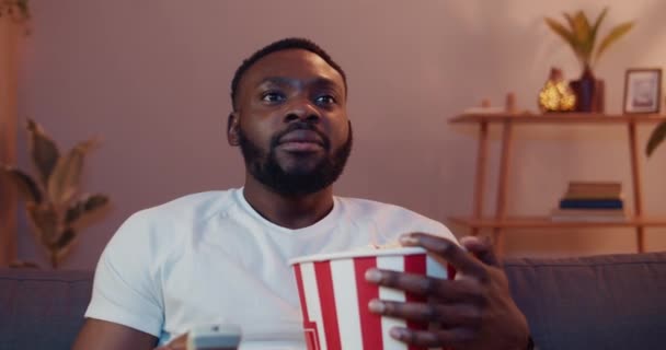 Front view of good looking guy switching on TV and watching television program. Young african man eating popcorn from paper bowl and using remote control while relaxing at home. - Video