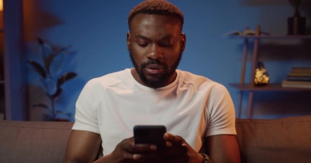 Front view of bearded african man in white t shirt typing message while sitting on sofa. Handsome guy using phone for communication in social networks while spending time at home. - Footage, Video