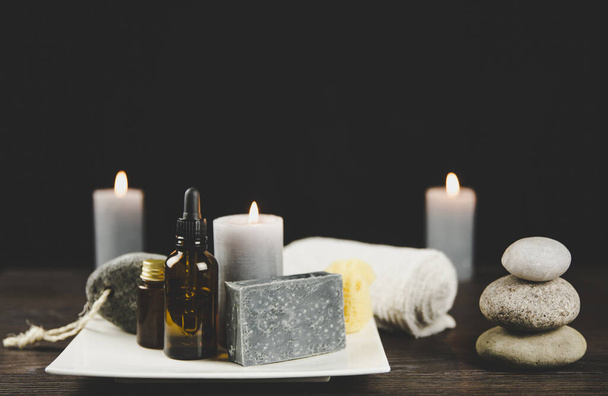 Men`s spa relaxation concept. Different day spa products( soap bar, beard oil, candles burning, rolled towel) on white ceramic tray on wooden table, dark black background. - Photo, Image