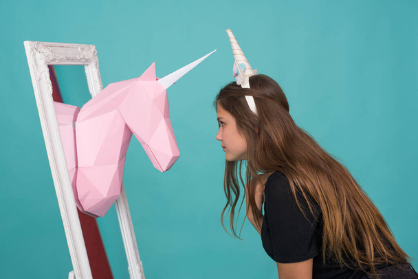 Adolescente Kawaii. Linda mujer joven con cuerno de unicornio mirando en el espejo y ver papel hecho a mano unicornio rosa sobre fondo azul
. - Foto, imagen