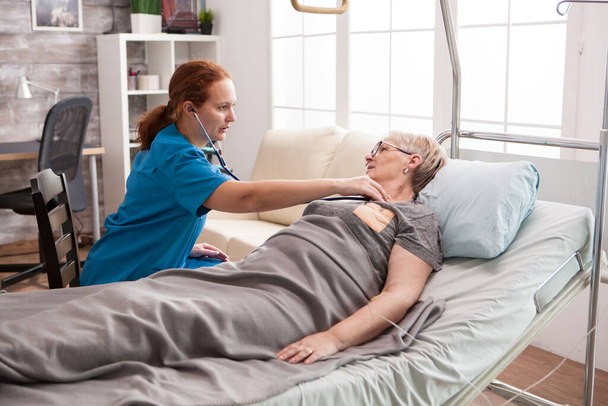 Молодая женщина-врач слушает сердцебиение старухи, лежащей в постели
 - Фото, изображение