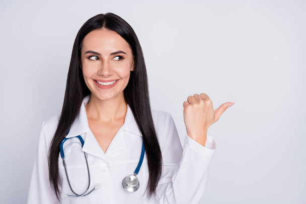 Photo of attractive beautiful doc lady good mood smiling patients direct thumb finger empty space insurance offer wear white lab coat stethoscope isolated grey color background - Fotoğraf, Görsel