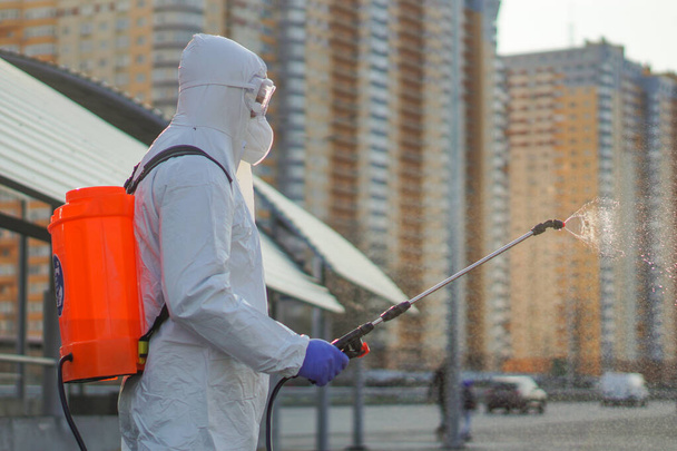 Koruyucu ekipman kullanan bir adam şehirde dezenfektanla dezenfekte ediyor. Coronavirus covid-19 hastalığından dolayı yüzey tedavisi. Beyaz takım elbiseli bir adam elinde sprey silahla caddeyi dezenfekte ediyor. Virüs salgını - Fotoğraf, Görsel