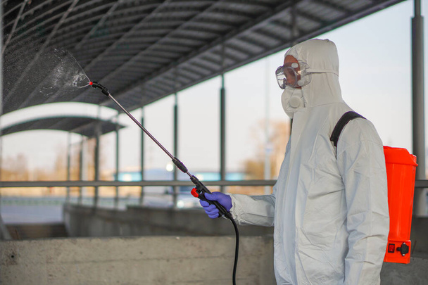 Een man in beschermende apparatuur desinfecteert met een sproeier in de stad. Oppervlaktebehandeling als gevolg van coronavirus covid-19 ziekte. Een man in een wit pak desinfecteert de straat met een spuitpistool. Viruspandemie - Foto, afbeelding