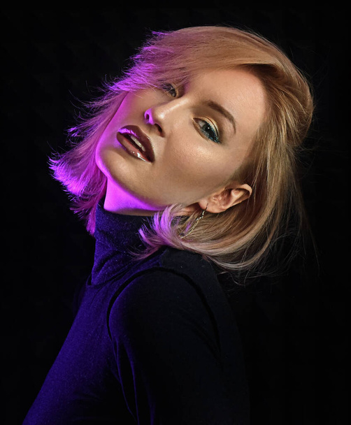 Sexy junge Frau posiert auf dunklem Hintergrund mit Neonlicht. Blondes Mädchen mit rosa Haaren porträtiert als Model für ein Style- Make-up Magazin. Leidenschaftliches Gesicht einer blonden Frau in einem dunklen Kleid, die glücklich aussieht - Foto, Bild