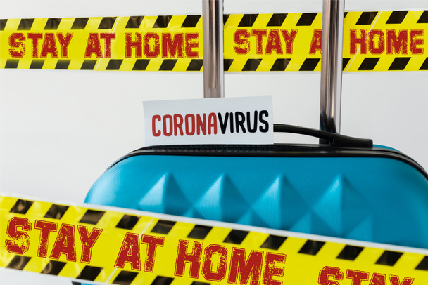 Nahaufnahme eines blauen Koffers mit Coronavirus-Karte in gelb-schwarzem Gefahrenwarnband mit zu Hause bleibender Abbildung isoliert auf weiß - Foto, Bild