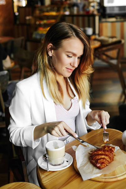 Beautiful young girl in a white jacket cuts a croissant sitting at a table in a cafe. Business lunch of a young girl. - Foto, Imagem