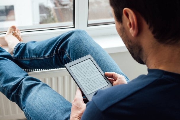 Sopra la spalla vista dell'uomo rilassante in jeans blu che legge un libro elettronico sul dispositivo digitale vicino alla finestra con i piedi nudi sdraiato su un davanzale di una finestra in un luminoso appartamento moderno
 - Foto, immagini