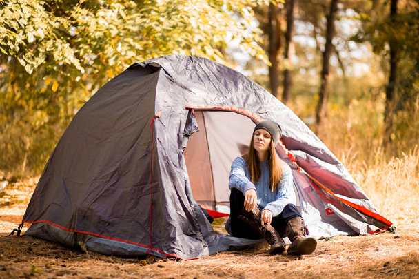 青いセーターと灰色の帽子でテントの近くに座っている若い女性観光客と秋の森の夕日で自由を楽しむ - 写真・画像
