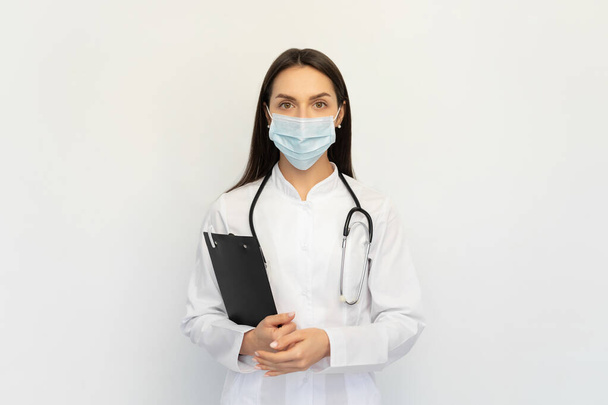 Medico femminile amichevole in una maschera medica protettiva con uno stetoscopio, tiene una cartella tra le mani
, - Foto, immagini
