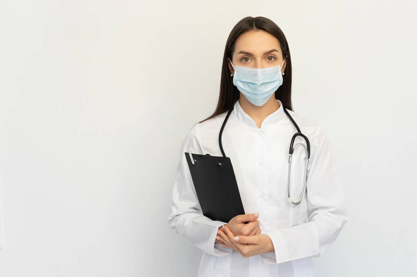 Medico femminile amichevole in una maschera medica protettiva con uno stetoscopio, tiene una cartella tra le mani
, - Foto, immagini
