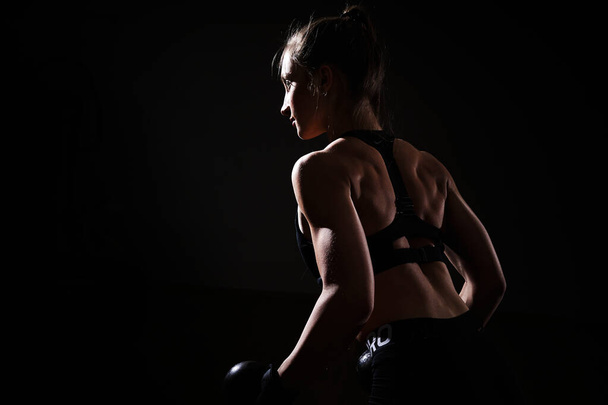 Νεαρή γυναίκα bodybuilder εργάζονται σε μαύρο φόντο. Γυναίκα με μυϊκό σώμα. - Φωτογραφία, εικόνα
