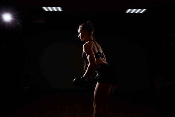 Beautiful girl athlete bodybuilder does exercises on dark background. Concept - the power of beauty, diet, sports. - Zdjęcie, obraz