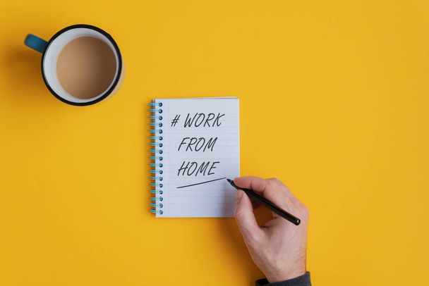 Männliche Hand schreibt ein Hashtag Work from home-Zeichen in einen Notizblock mit einer Tasse Tee oder Kaffee daneben. Vor gelbem Hintergrund. - Foto, Bild