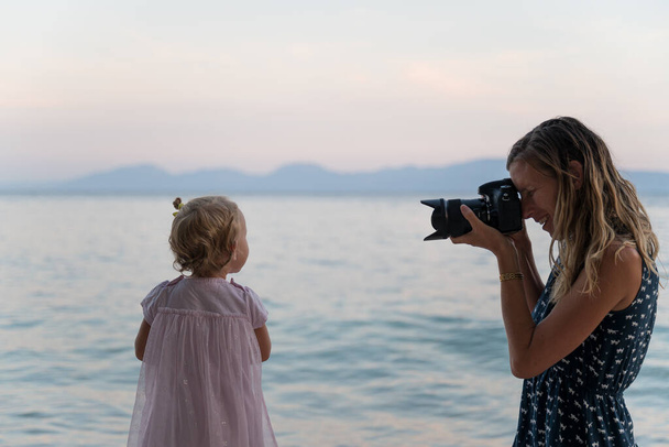 Sorridente giovane madre scattare una foto della sua figlia bambino in piedi sulla spiaggia proprio vicino al mare serale
. - Foto, immagini