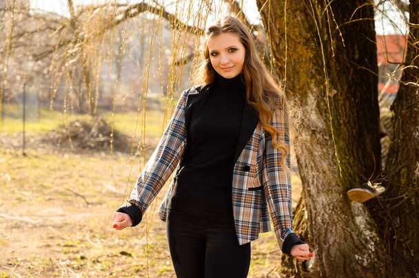 Spring portrait of a fashionable beautiful young girl with long hair with a smile in the park on a wood background. Photo taken in sunny weather outdoors. - Fotografie, Obrázek