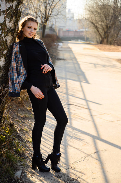 Full-length portrait of a stylish young woman with long hair with a smile in the city against the background of a wall of a business building with windows. Photo in sunny weather outdoors in spring. - Fotoğraf, Görsel