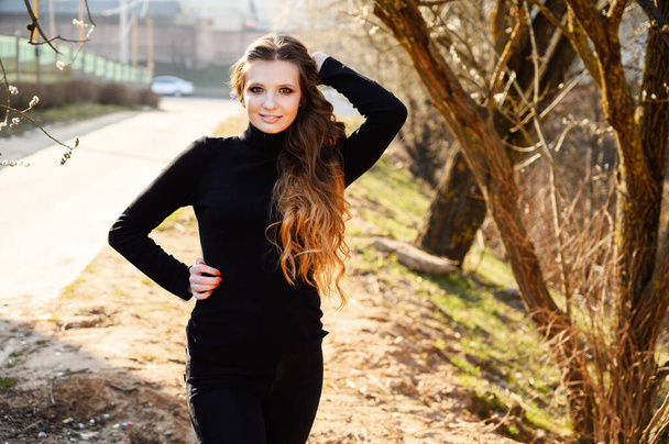 Portrait of a stylish young woman with long hair with a smile on the background of road trees in the park. Photo in sunny weather outdoors in spring. - Fotografie, Obrázek
