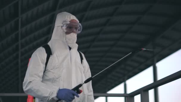 Een man in beschermende apparatuur desinfecteert met een sproeier in de stad. Oppervlaktebehandeling als gevolg van coronavirus covid-19 ziekte. Een man in een wit pak desinfecteert de straat met een spuitpistool. Viruspandemie - Video