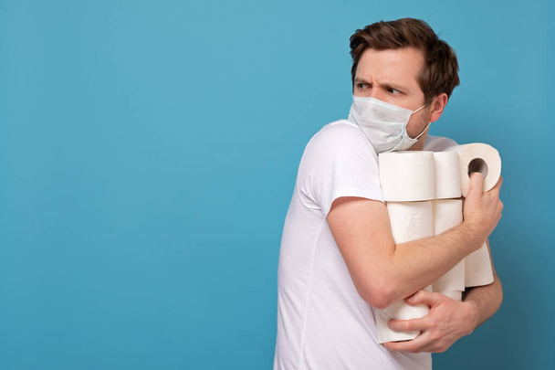 Chciwość i panika z powodu koronawirusu. Mężczyzna w masce medycznej, trzymający dużo rolek papieru toaletowego, ukrywający je przed wszystkimi. Studio zdjęcie na niebieskiej ścianie - Zdjęcie, obraz