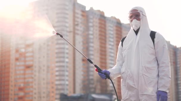 Koruyucu ekipman kullanan bir adam şehirde dezenfektanla dezenfekte ediyor. Coronavirus covid-19 hastalığından dolayı yüzey tedavisi. Beyaz takım elbiseli bir adam elinde sprey silahla caddeyi dezenfekte ediyor. Virüs salgını - Video, Çekim