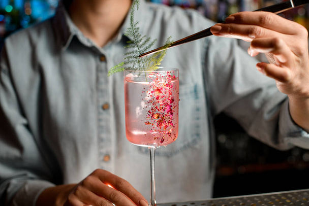 Крупным планом молодая женщина-бармен украшает стекло розовым коктейлем за куском зеленого растения
. - Фото, изображение