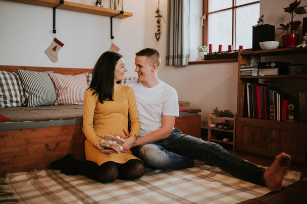 Young pregnant woman and man with baby boots. Loving young couple expecting baby sitting on floor and smiling. - Foto, Bild