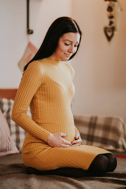 Pregnant young woman touching baby bump. Young woman expecting baby kneeling on bed touching tummy and smiling. - Fotoğraf, Görsel