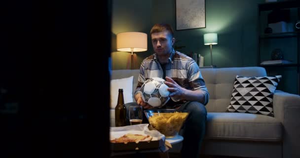 Jóképű fehér férfi focirajongó ül a kanapén és késő este focimeccset néz. Guy éljenzés és aggódás kedvenc csapat előtt TV sport csatorna és bajnokság játék. - Felvétel, videó