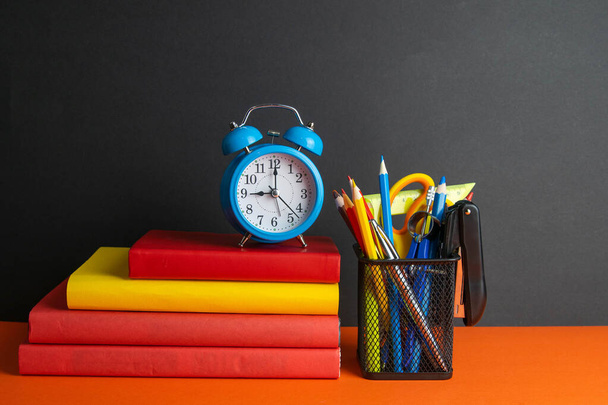 A stack of books, a green flower, blue clock and a glass of pencils stand on an orange background. - Фото, изображение