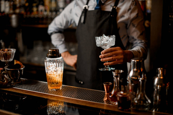 barman holds glass with ice. Glassy cocktail shaker stands on bar counter. - 写真・画像