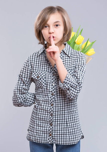 Girl hiding bunch of flowers behind itself, on gray background. Child with bouquet of yellow tulips putting finger up to lips and saying shhh. Happy mothers, Birthday or Valentines day. - 写真・画像