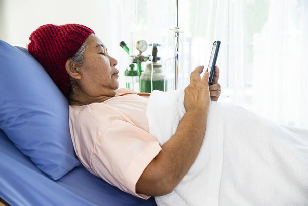 Asian elderly female wearing wool hat used smartphone while lying on patient bed in hospital - Fotó, kép